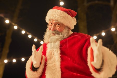 Téléchargez les photos : Portrait taille haute du Père Noël souriant invitant les gens tout en restant debout à l'extérieur - en image libre de droit