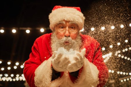 Foto de Retrato de Papá Noel tradicional soplando nieve a la cámara al aire libre con luces de hadas en el fondo - Imagen libre de derechos