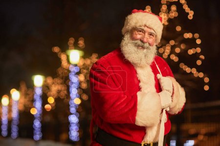 Photo for Waist up portrait of traditional Santa Claus holding sack with presents and smiling at camera in night city with lights, copy space - Royalty Free Image