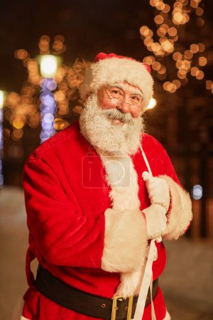 Foto de Retrato vertical de Papá Noel tradicional sosteniendo saco con regalos y sonriendo a la cámara en la ciudad nocturna - Imagen libre de derechos