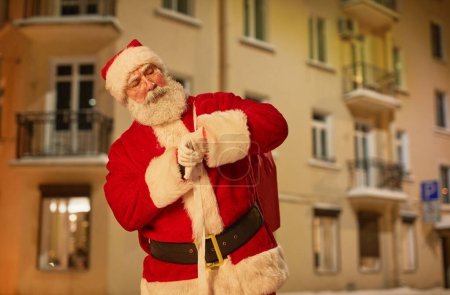Téléchargez les photos : Portrait taille haute du Père Noël traditionnel vérifiant l'heure sur montre-bracelet tout en se tenant debout à l'extérieur la veille de Noël, espace de copie - en image libre de droit