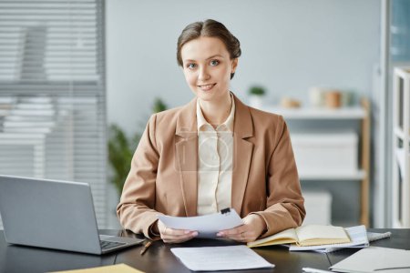 Foto de Retrato mínimo de una joven empresaria sonriendo a la cámara mientras está sentada en el escritorio en la oficina y sosteniendo contratos - Imagen libre de derechos