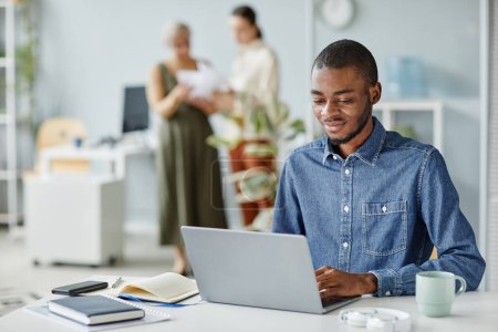 Téléchargez les photos : Portrait de jeune homme noir utilisant un ordinateur portable au bureau dans un espace intérieur minimal, espace de copie - en image libre de droit