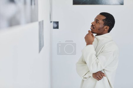 Téléchargez les photos : Vue de côté portrait de bel homme noir regardant des images dans la galerie de photos et appréciant l'art, l'espace de copie - en image libre de droit