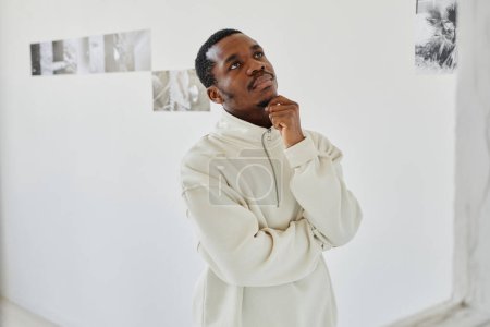 Téléchargez les photos : Portrait taille haute de jeune homme afro-américain vêtu de blanc regardant des photos dans la galerie d'art, espace de copie - en image libre de droit