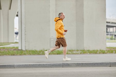 Photo for Side view full length portrait of handsome mature man running outdoors in city against concrete background, copy space - Royalty Free Image