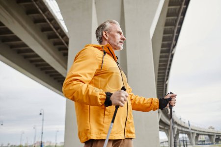 Photo for Side view portrait of sportive mature man enjoying nordic walk with poles in urban city setting, copy space - Royalty Free Image