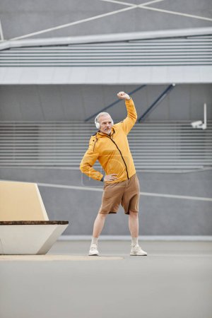 Téléchargez les photos : Portrait vertical complet de l'homme mûr sportif s'étirant à l'extérieur avec écouteurs dans un cadre urbain - en image libre de droit