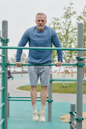 Photo for Full length portrait of sportive mature man exercising on parallel bars outdoors and looking at camera - Royalty Free Image