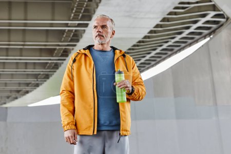Téléchargez les photos : Portrait taille haute d'un homme âgé actif tenant une bouteille d'eau sur fond de béton en milieu urbain, espace de copie - en image libre de droit