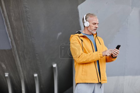 Foto de Retrato de vista lateral del hombre mayor guapo con auriculares al aire libre y la creación de música a través de teléfono inteligente contra el fondo de hormigón, espacio de copia - Imagen libre de derechos