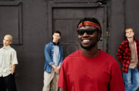 Foto de Diverso grupo de niños que usan ropa de estilo callejero de pie contra la pared negra al aire libre y mirando a la cámara, se centran en sonreír hombre afroamericano - Imagen libre de derechos