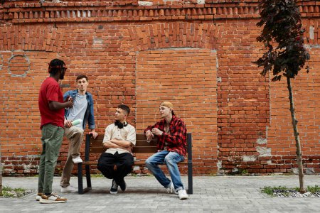 Téléchargez les photos : Vue grand angle à divers groupes de jeunes hommes portant la mode de style de rue dans la zone urbaine de la ville contre le mur de briques et bavarder, copier l'espace - en image libre de droit