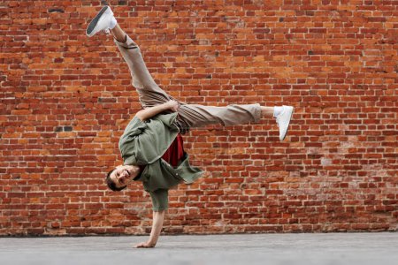 Téléchargez les photos : Prise de vue complète du jeune homme faisant la pose du handstand hip-hop et souriant à la caméra contre un mur de briques, espace de copie - en image libre de droit