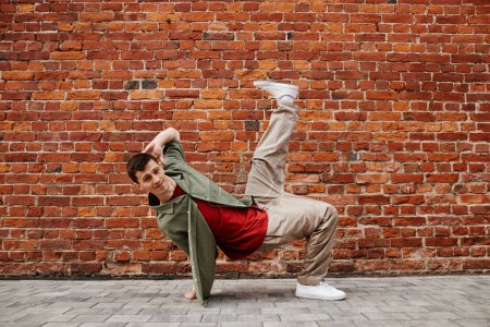 Foto de Largura completa de disparo de hombre joven haciendo hip-hop pose de baile y sonriendo a la cámara contra la pared de ladrillo, espacio de copia - Imagen libre de derechos