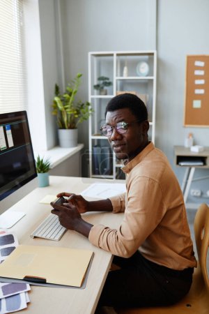 Foto de Retrato vertical del joven negro mirando la cámara en el lugar de trabajo de la oficina mientras diseña la interfaz de la aplicación móvil - Imagen libre de derechos
