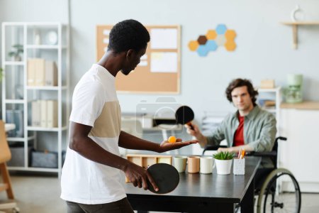 Foto de Retrato de un joven negro jugando al tenis de mesa en la oficina contra un colega con discapacidad, concepto de inclusión en el lugar de trabajo - Imagen libre de derechos