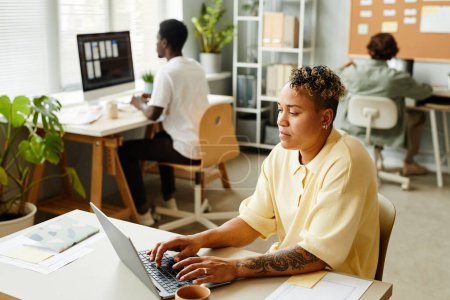 Téléchargez les photos : Portrait de femme noire tatouée utilisant un ordinateur portable au bureau tout en travaillant avec l'équipe en arrière-plan, espace de copie - en image libre de droit