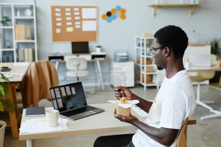 Téléchargez les photos : Portrait latéral de jeune homme noir mangeant des nouilles à emporter au travail pendant la pause déjeuner, espace de copie - en image libre de droit