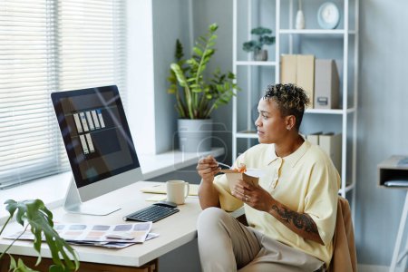 Téléchargez les photos : Portrait latéral de femme noire tatouée mangeant des plats à emporter sur le lieu de travail pendant la pause déjeuner et regardant l'écran d'ordinateur - en image libre de droit