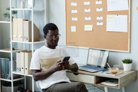Photo for Portrait of young black man using smartphone at workplace in office and texting or scrolling social media during break time - Royalty Free Image