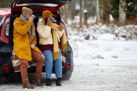 Photo for Full length portrait of young couple sitting in car trunk in winter forest while enjoying hot cocoa, copy space - Royalty Free Image
