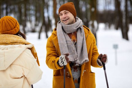Photo for Waist up portrait of happy couple skiing in winter forest, focus on carefree young man with ski poles smiling at girlfriend - Royalty Free Image