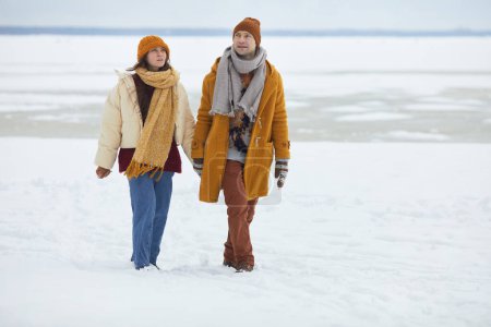 Foto de Retrato de larga duración de una pareja joven cogida de la mano y caminando hacia la cámara en un paisaje invernal mínimo, espacio para copiar - Imagen libre de derechos