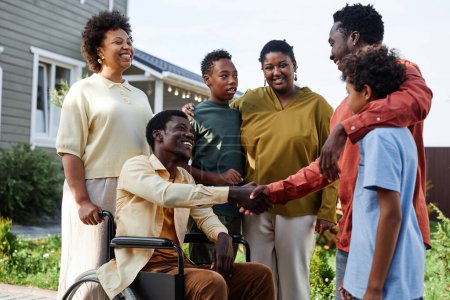 Foto de Retrato de vista lateral de la gran familia afroamericana con persona en silla de ruedas dando la bienvenida a los invitados para la fiesta de verano - Imagen libre de derechos