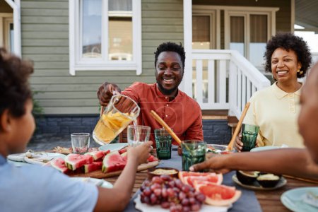 Foto de Retrato del hombre negro sonriente vertiendo jugo de naranja al vidrio durante la reunión familiar al aire libre - Imagen libre de derechos