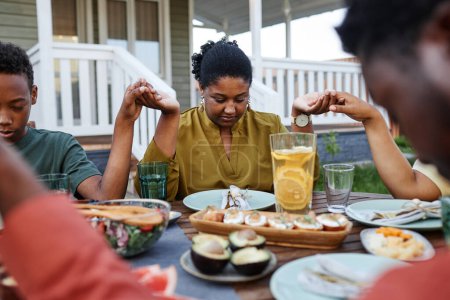 Téléchargez les photos : Portrait de jeune femme afro-américaine disant grâce à table à l'extérieur pendant la réunion de famille et se tenant la main - en image libre de droit