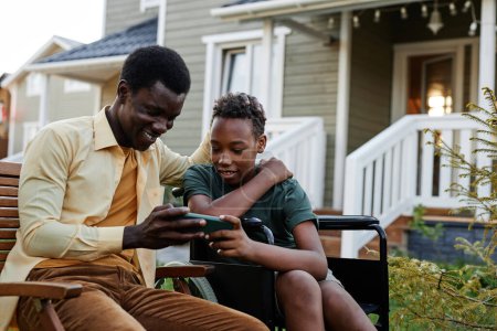 Téléchargez les photos : Portrait d'un adolescent souriant regardant l'écran du smartphone tout en passant du temps avec son père à l'extérieur - en image libre de droit