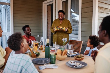Téléchargez les photos : Portrait de femme noire souriante apportant de la nourriture à table tout en appréciant la réunion de famille à l'extérieur - en image libre de droit