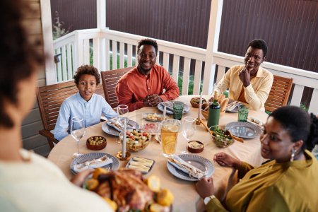 Foto de Vista de ángulo alto de la familia afroamericana feliz en la mesa de la cena al aire libre con la mujer que trae platos caseros - Imagen libre de derechos