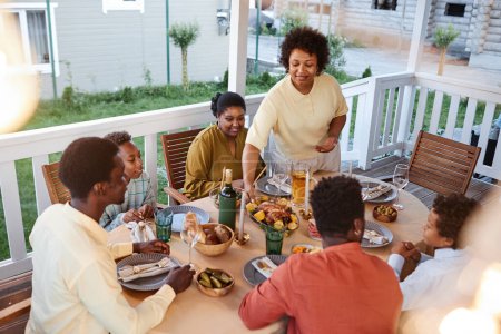 Foto de Familia afroamericana en la mesa al aire libre con una mujer sonriente que trae platos caseros - Imagen libre de derechos