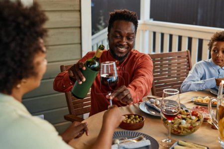 Foto de Retrato de hombre negro sonriente vertiendo vino a las copas mientras disfruta de la cena al aire libre - Imagen libre de derechos