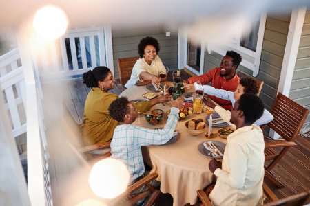 Foto de Vista de ángulo alto en grandes vasos de tintineo de la familia afroamericana sobre la mesa de la cena al aire libre en un ambiente acogedor con luces de hadas - Imagen libre de derechos
