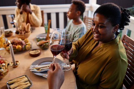 Téléchargez les photos : Vue de côté portrait de souriante femme noire cliquetis verres à vin à la table du dîner à l'extérieur dans un cadre confortable - en image libre de droit