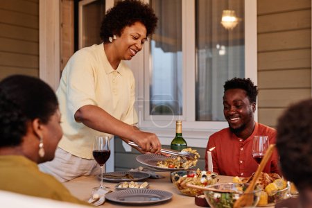 Téléchargez les photos : Portrait d'une femme afro-américaine souriante servant de la nourriture à sa famille tout en dînant ensemble à l'extérieur le soir - en image libre de droit