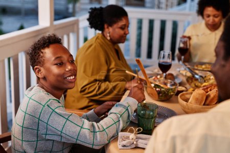 Téléchargez les photos : Portrait latéral d'un garçon afro-américain souriant profitant d'un dîner en famille sur la terrasse à l'extérieur - en image libre de droit