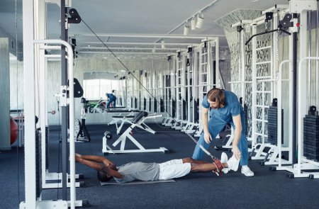 Photo for African young patient lying on floor and doing physical exercise on his leg with the help of doctor in gym - Royalty Free Image