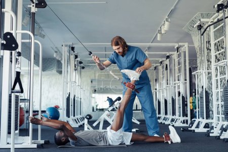Téléchargez les photos : Médecin masculin en uniforme aidant le patient handicapé à étirer sa jambe avec un équipement sportif spécial - en image libre de droit