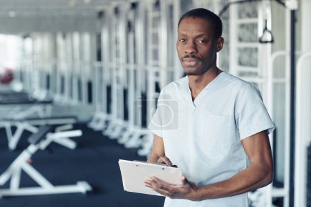 Photo for Portrait of African doctor in uniform filling patient medical card while standing in gym - Royalty Free Image