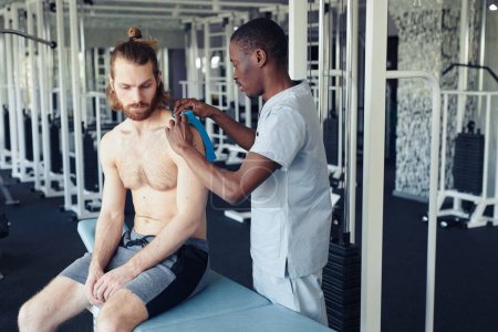 Téléchargez les photos : Jeune médecin africain mettant des bandes sur les ligaments du patient avant l'entraînement en salle de gym - en image libre de droit