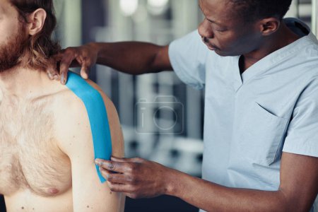 Téléchargez les photos : Thérapeute africain appliquant de la bande sur l'épaule du patient avant les exercices physiques dans la salle de gym - en image libre de droit