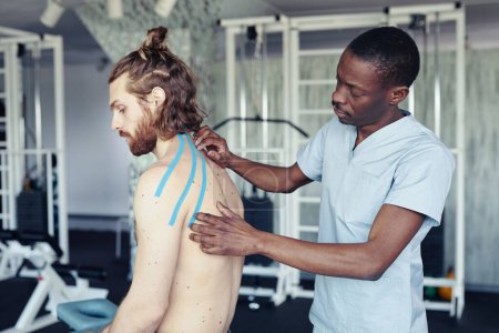 Téléchargez les photos : Jeune thérapeute africain appliquant des bandes sur le dos du patient pour traiter les ligaments avant l'entraînement sportif - en image libre de droit