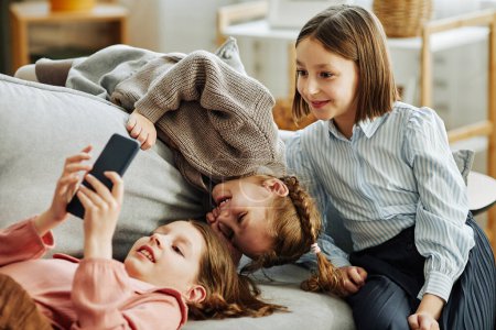 Foto de Retrato de tres niñas jugando juntas en el sofá en casa y usando un teléfono inteligente - Imagen libre de derechos