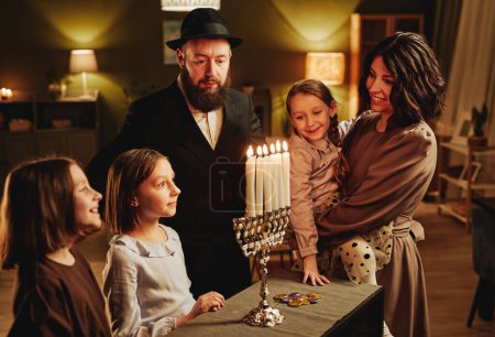 Foto de Retrato de la familia judía moderna mirando la vela de plata menorah durante la celebración de Hanukkah en el acogedor hogar - Imagen libre de derechos