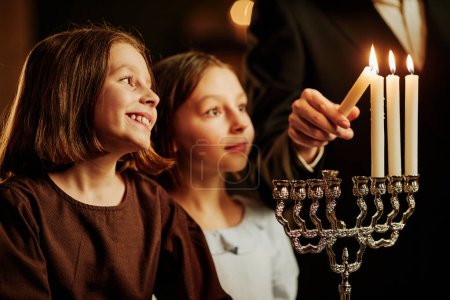 Foto de Retrato de vista lateral de dos niñas judías mirando la vela Menorah y sonriendo felizmente durante la celebración de Hanukkah, espacio para copiar - Imagen libre de derechos