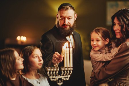 Portrait of orthodox jewish man lighting menorah candle with family during Hanukkah celebration